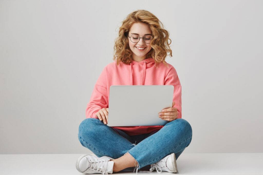 smiling female student writing essay sitting with laptop floor