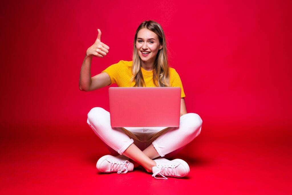 happy young curly beautiful woman sitting floor with crossed legs using laptop red wall